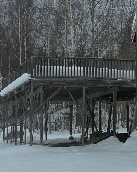 Altan med trästaket och snö på taket, stöttad av lutande pelare i vintrigt skogslandskap.