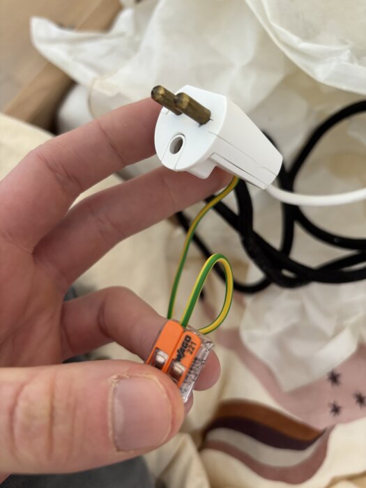 Hand håller i en jordad lamppropp och WAGO-koppling med gulgrön kabel mot en oskarp bakgrund.