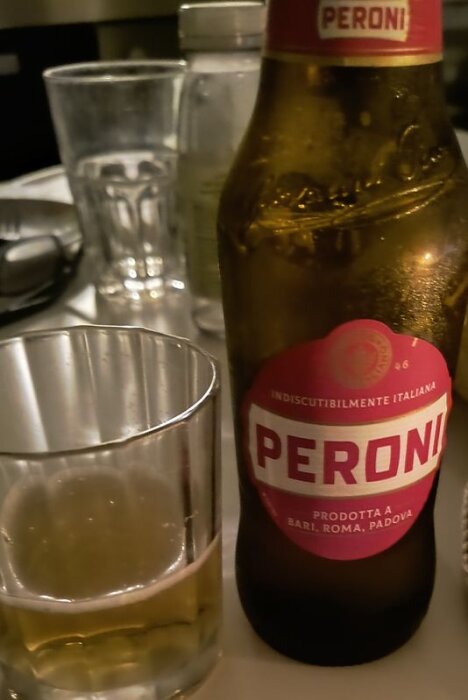 En flaska Peroni öl står på ett bord bredvid ett halvt fyllt glas med öl, andra glas och föremål i bakgrunden.
