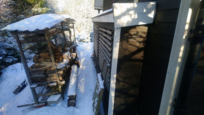 Snöig “låda” med vedstapel vid husvägg, delvis skuggad, synlig kofångare från en Amazon, vintermiljö med snö.
