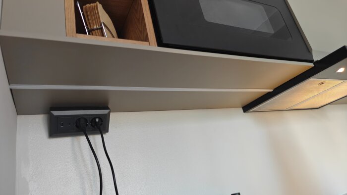 Väggmonterat eluttag med två sladdar, placerat under ett hyllplan i köket med inbyggd LED-belysning och fläkt.