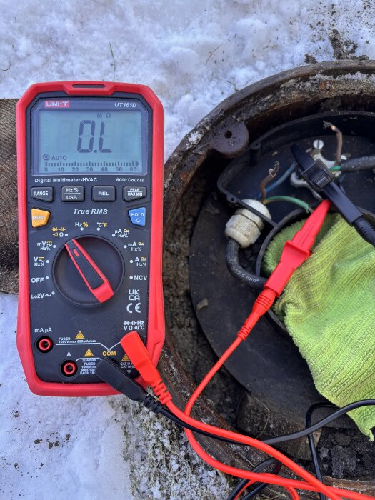 Digital multimeter vid pumphål, visar 0 L ohm i snöigt område, med kablar anslutna och en arbetsduk.