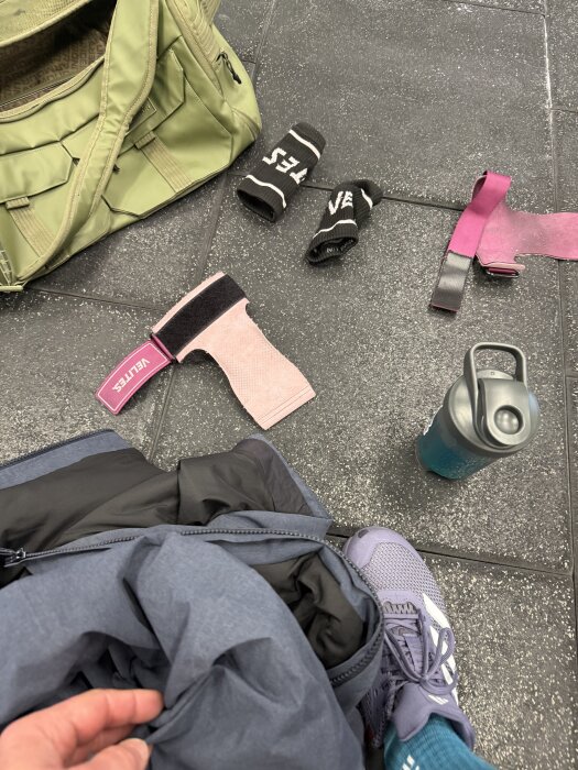 En gymväska ligger på golvet med träningshandskar, en vattenflaska och sneakers. En hand håller i en jacka.