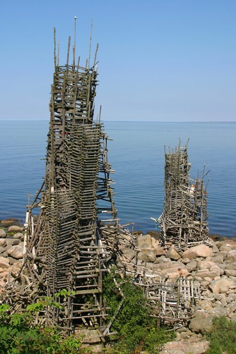 Tornliknande skulpturer av träpinnar vid kusten, som påminner om Nimis av Lars Vilks, med en stenig strand i bakgrunden.