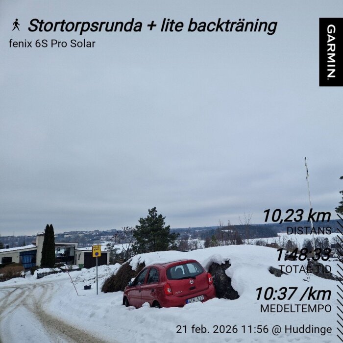 En röd bil fast i snö vid sidan av en väg; landskap med snötäckt mark och utsikt över tak i bakgrunden.