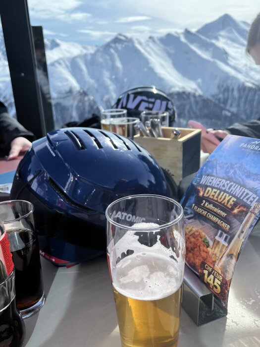 Ölglas och skidutrustning på ett bord med alplandskap i bakgrunden. Meny med schnitzelreklam synlig.