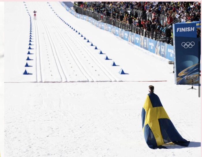 Skidåkare klädd i svensk flagga står vid mållinjen och väntar på en konkurrent som närmar sig i bakgrunden under ett skidlopp.