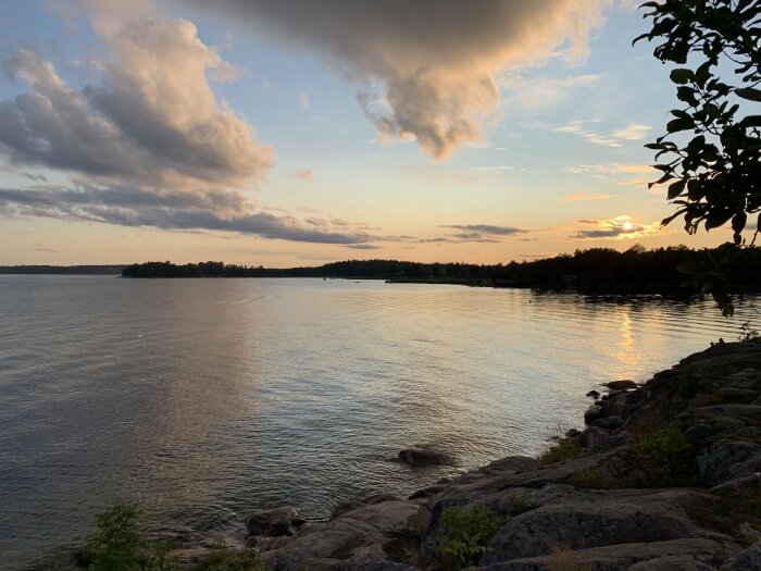Solnedgång över lugn havsvik med molnig himmel, klippa i förgrunden.