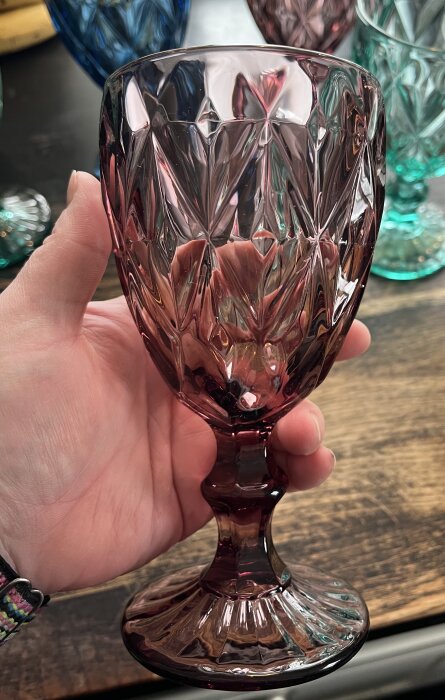 En person håller ett färgglatt, mönstrat vinglas mot en träyta med fler glas i bakgrunden.