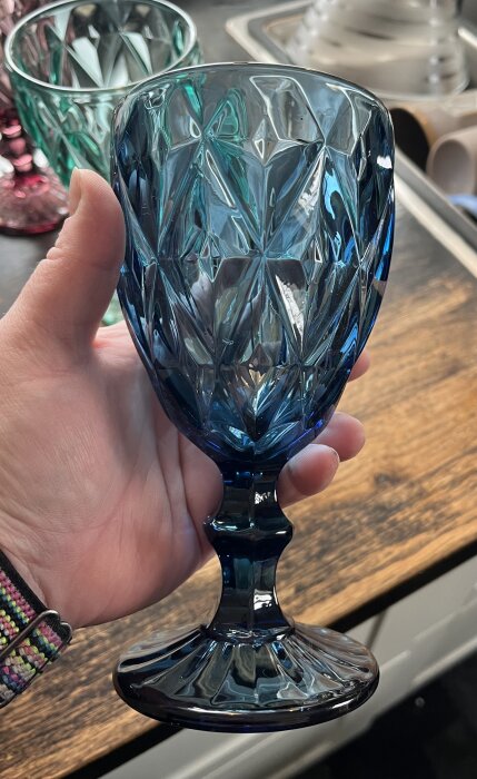 En hand håller ett blått, facetterat vinglas i glas med färgglada glas i bakgrunden.