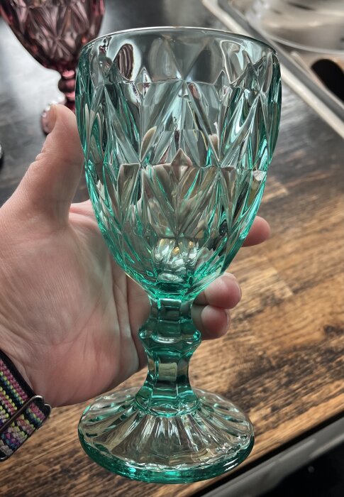 En hand håller i ett turkost kristallglas med detaljerade mönster, står på ett träbord. Bakgrunden har ett rosa glas.