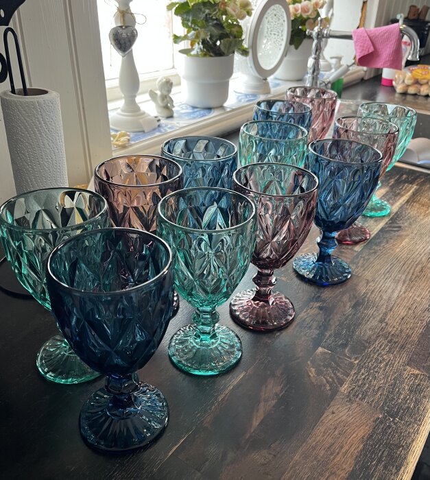 Flera färgglada och mönstrade glas står uppradade på ett köksbord i ett soligt rum med växter och inredningsdetaljer i bakgrunden.