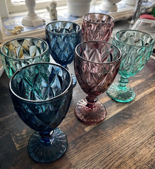 Sex färgglada kristallglas på ett bord, i olika nyanser av blått, grönt och rosa, med en dekorativ, slipad design.