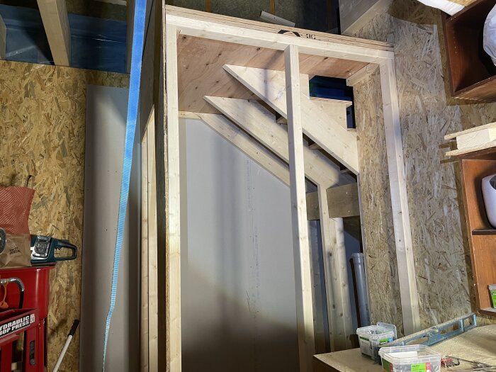 Rå struktur av en toalett under konstruktion i ett garage, med synliga träbjälkar och plywoodskivor. Vinklade vangstycken i trappan.