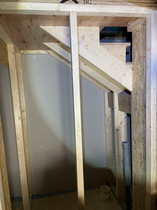 Toalettkonstruktion i garage med träram och synliga vangstycken för trappa. Dubbla plywoodskivor används för golv och tak.