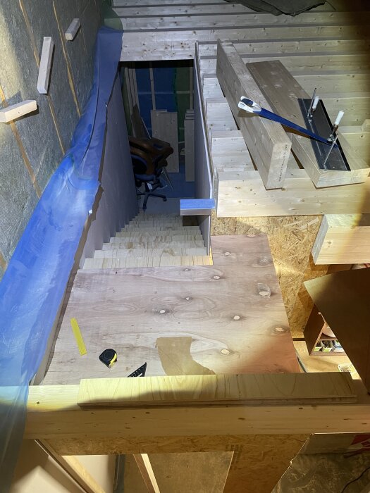 Trappa under konstruktion i ett garage, med plywoodgolv och byggmaterial som tumstock och måttband; vangstycken och takbjälkar syns.