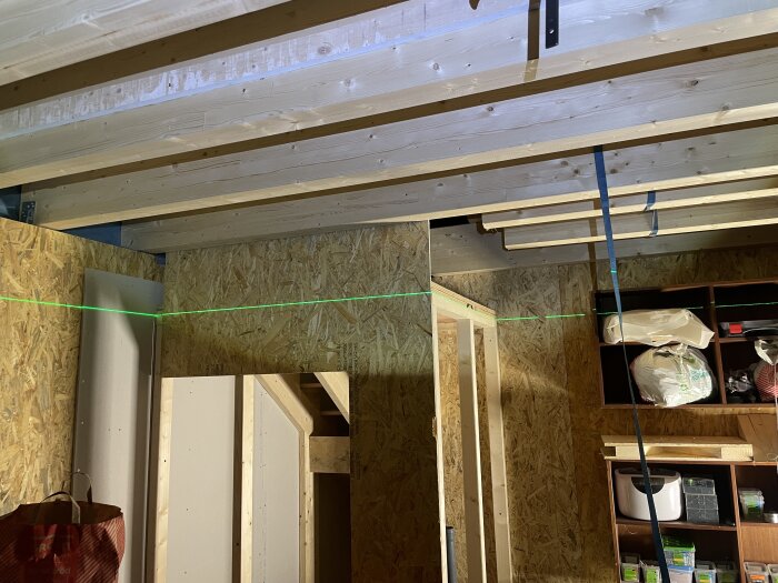 Byggprojekt med träväggar och takbjälkar, där en grön laserlinje visar rätt höjd. Hyllor med verktyg och material finns i bakgrunden.