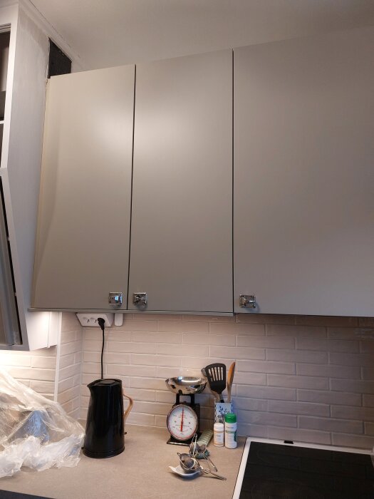 Moderna köksskåp med köksredskap på bänken, ett eluttag och köksfläkt gömd bakom skåp i en flerfamiljslägenhet med självdragssystem.