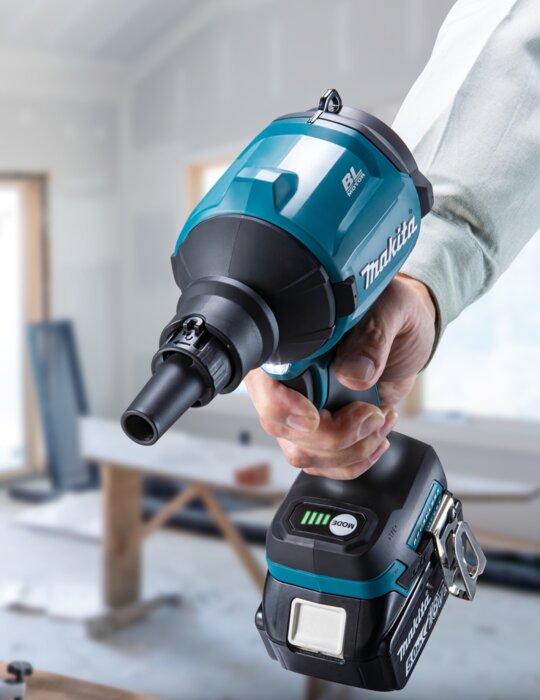 En person håller en blå och svart Makita-blåsapparat med batteri i ett rum under renovering.