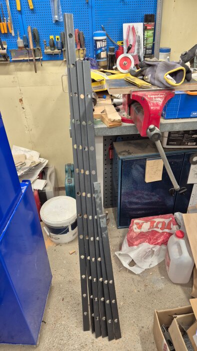 Svarta metallstänger med många förborrade hål, stående lutade mot en arbetsbänk i en verkstad full av verktyg och material.