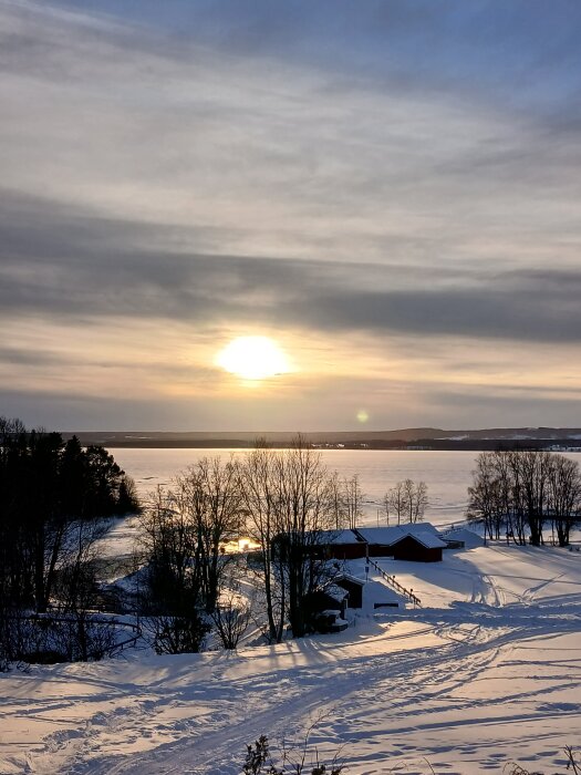 Solnedgång över en snötäckt landsbygd med träd och röda byggnader vid en frusen sjö.