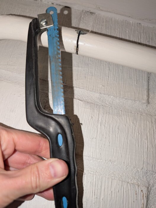 Hand håller bågsåg mot vit plastledning, linje markerad för kapning.