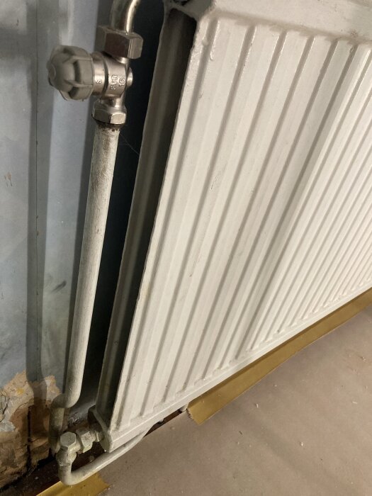 Gammaldags metallradiator med anslutna rör och ventil, monterad mot en vägg med sliten färg.