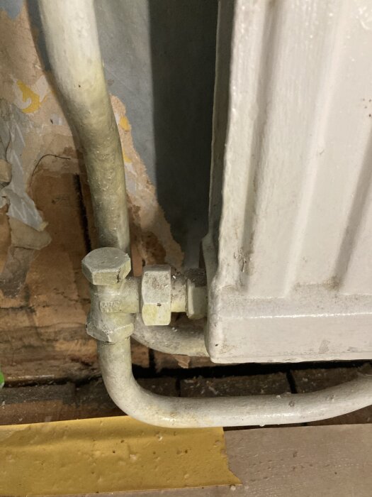 Radiator med kopplingar och rör, synliga mot en vägg med sliten tapet och gammal tretex.