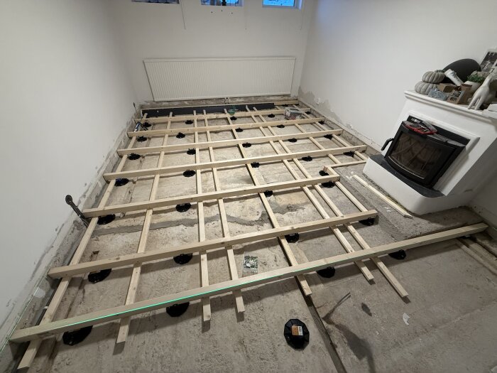 Träram för golvkonstruktion på betonggolv intill en kamin, förberedd för installation av ett flytande golv i ett rum med vita väggar.