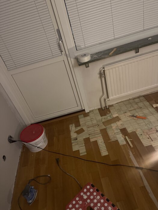Rum med trasigt trägolv, delvis avtäckt med verktyg runtom, bredvid balkongdörr och element. En burk och elkabel syns på golvet.