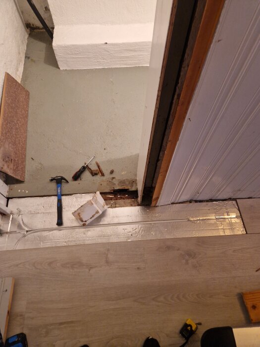 Bild på ett golvområde i ett källarutrymme, där en trasig träregel, golvverktyg och en avlägsnad isoleringsbit syns.