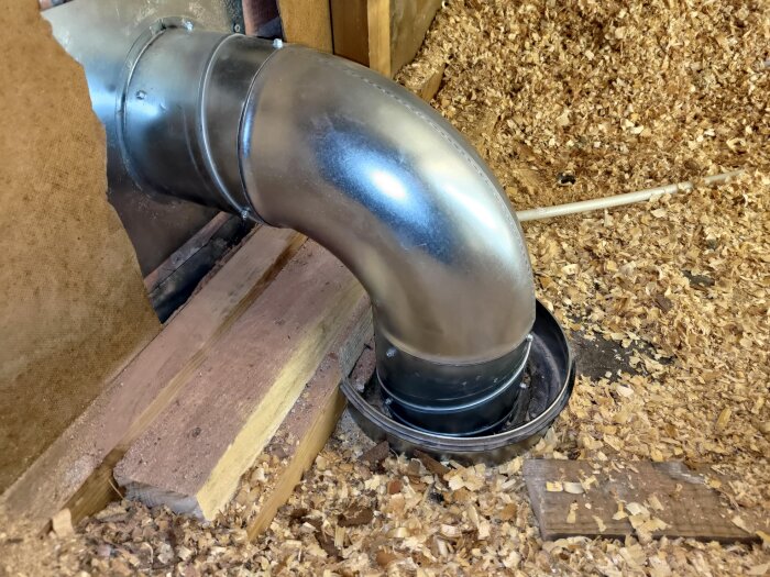 Metalliskt spirorör monterat genom vägg, omgivet av spån och träbitar, del av ett köksventilationssystem.