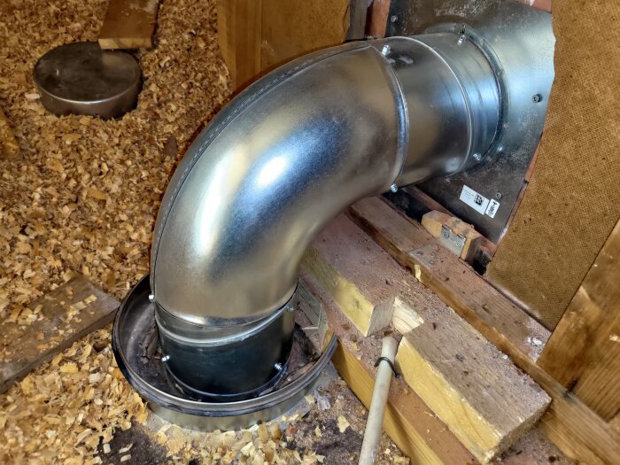 Spirorör på 160mm installerat genom yttervägg ovanför köksfläkt, omgiven av sågspån och fastsatt med skruvar.