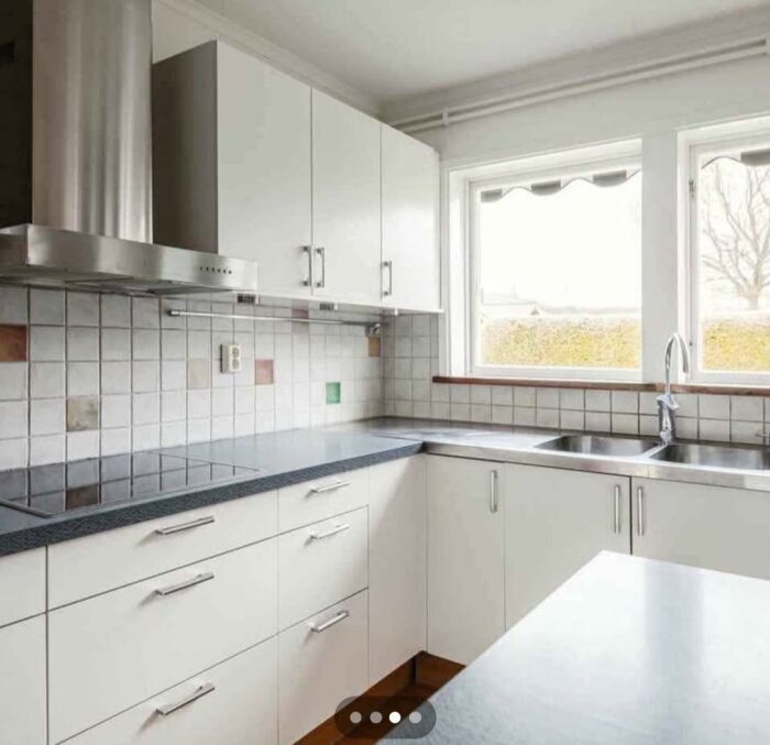 Ljust kök med vit köksinredning, hörnhäll, integrerad ugn och mikro samt rostfri fläkt. Två fönster ger naturligt ljus. Grått kakel ovanför diskbänken.