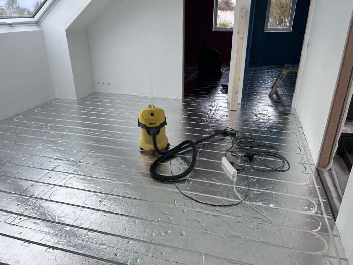 Golvet i ett rum med installerad golvvärme och en gul dammsugare på en plastig yta, trägolv och diverse verktyg synliga.