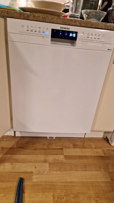 Vit Siemens iQ300 diskmaskin i ett kök, med trälaminatgolv och disk ovanpå bänkskivan. Digital display visar information om programtid.