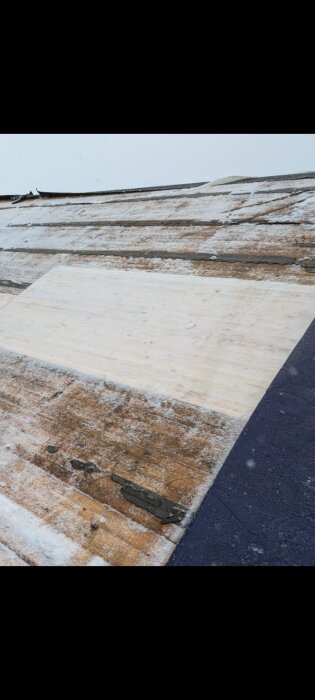 Snötäckt tak med slitna träplankor, delvis nya plywoodskivor, tecken på slitage och snö på ytan.