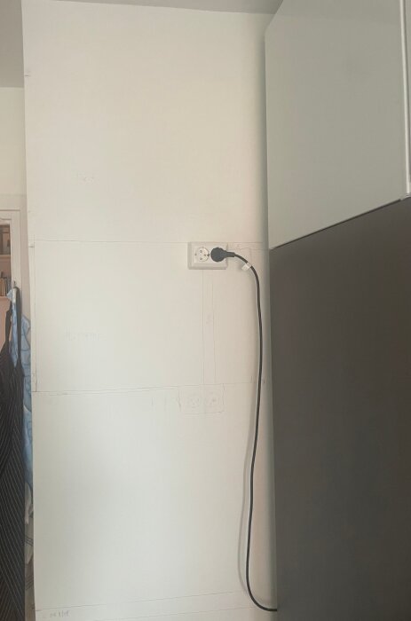 Vit vägg med skissade linjer och eluttag, kabel ansluten i uttaget, intill en köksskåp.