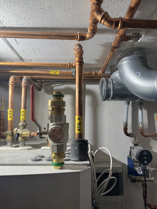 Kopparrör och ventilsystem i en källare, med märkta ledningar och en monterad pump på väggen.