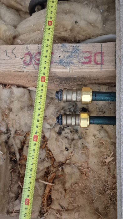 Två rör med mässingskopplingar ligger i isolering bredvid ett måttband som visar 98-112 cm, i ett delvis öppet byggprojekt.