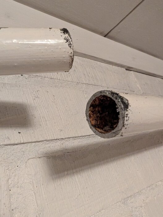 Närbild av ett avsågat vitt metallrör mot en vit träbakgrund. Rören är åtskilda och ena änden är korroderad.