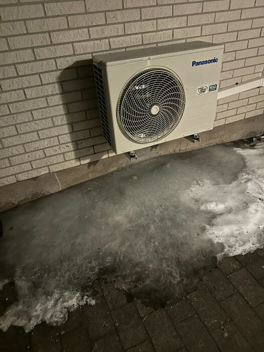 Utomhusenhet för värmepump ovanför en isig yta vid tegelvägg.