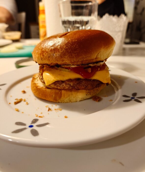 Hamburgare med ost och ketchup på ett mönstrat vitt fat, stående på ett dukat bord med glas i bakgrunden.