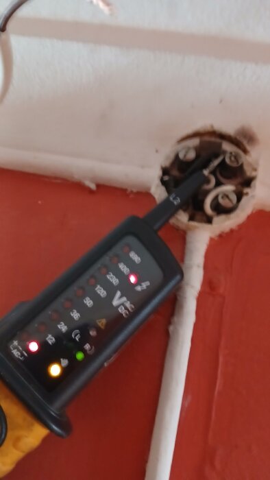 Spänningsmätare visar 230V i vägguttag med kopplingskablar i gammal installation.