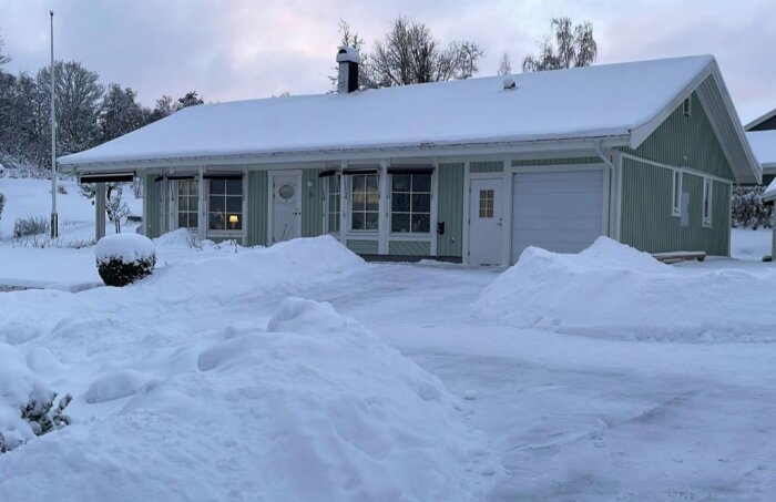 Framsida av ett grönmålat hus med snötäckt mark och uppfart, sett under en vinterdag.