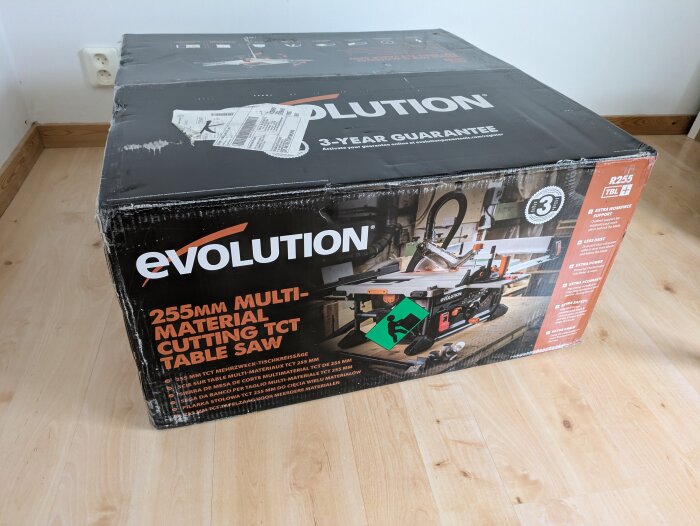 Kartong med texten "Evolution 255mm Multi-Material Cutting TCT Table Saw" på ett trägolv.