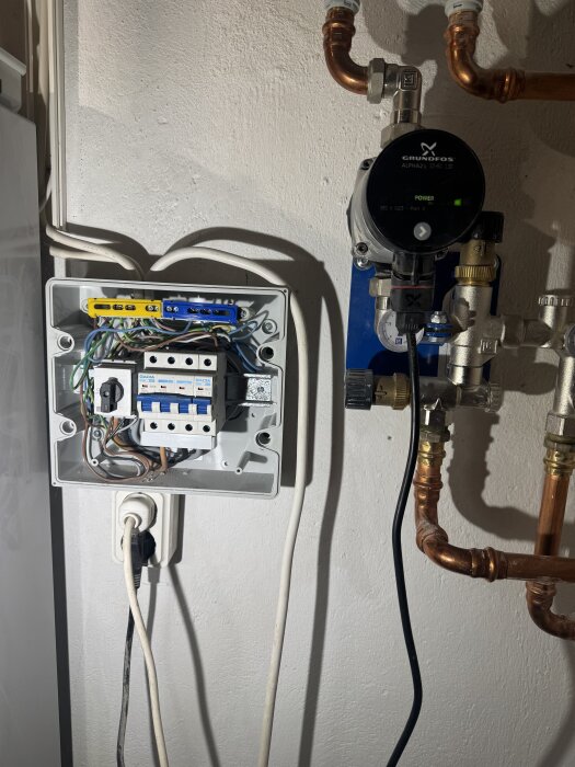 Öppen elcentral med säkringar på väggen bredvid en Grundfos pump och kopparrör i pannrum.