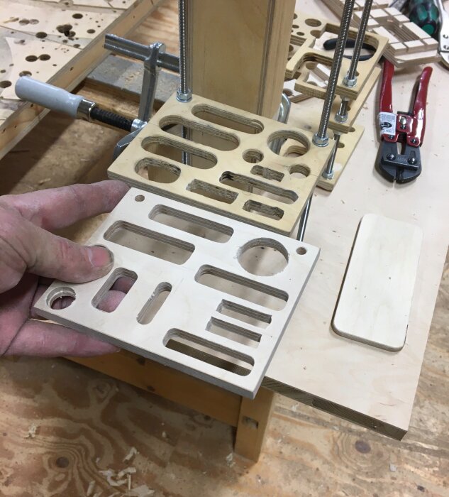 Hand håller en utskuren plywoodbit med olika former, omgiven av verktyg och träbearbetningsmaterial på ett arbetsbord.