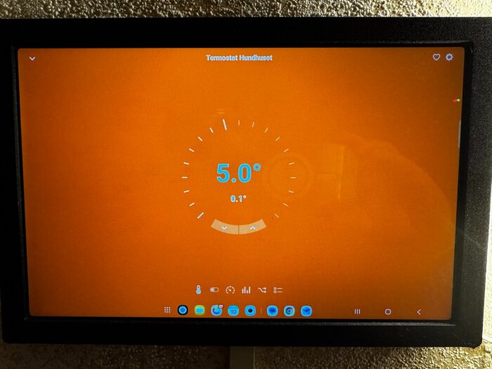 Digital termostatdisplay med orange bakgrund som visar 5.0 grader för "Termostat Hundhuset", med ikoner för inställningar nedtill.