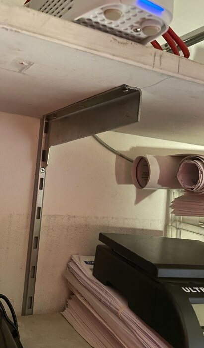 Hylla med metallkonsoll och väggskena i ett rum, en router ovanpå hyllan och papper staplade bredvid.
